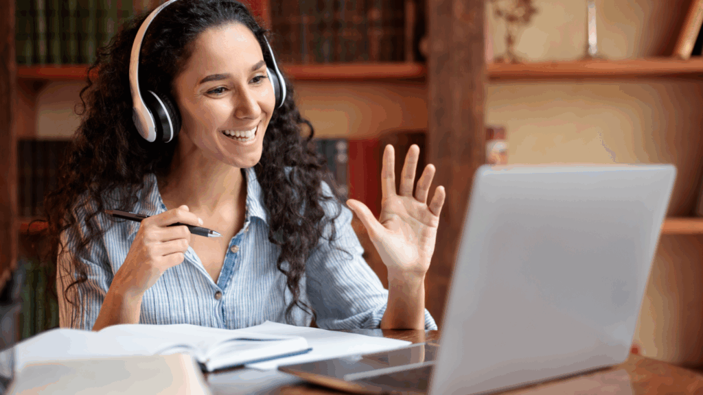 A smiling woman is wearing headphones and teaching online using a laptop.