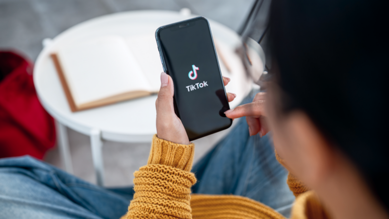 A person in a yellow sweater holds a smartphone displaying the TikTok app. An open book lies on a white table in the background.