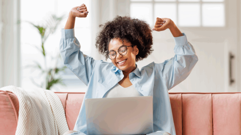 A joyful woman with curly hair and glasses raises her arms in celebration while sitting on a pink sofa, using a laptop.