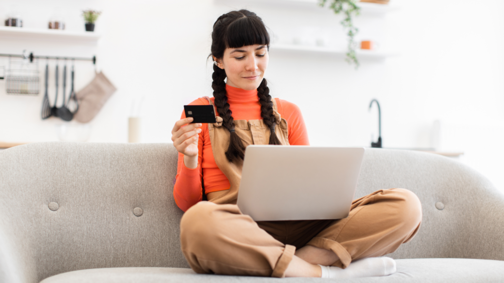 A woman sitting on top of a couch with a laptop on her lap, holding a debit card in her hand.
