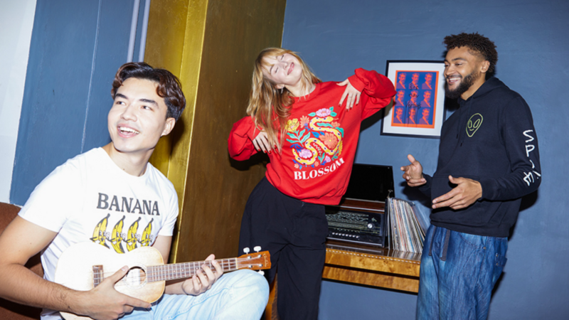 A group of young people (2 men and 1 woman) is happily dancing in a room, wearing stylish casual clothes.