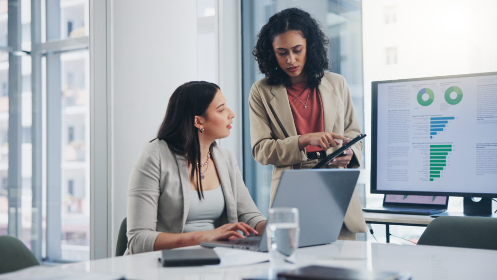 In an office, two women engage in discussion as they review information on a laptop.