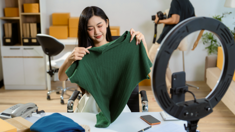 A woman smiles while holding a green shirt in front of a camera setup with a ring light, as if reviewing or showcasing the item.