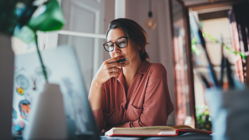 A woman holding a pen in her hand and looking at a laptop screen, thinking.