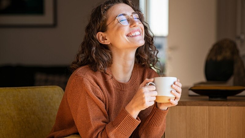 Smiling woman in glasses holding a white mug while relaxing at home.