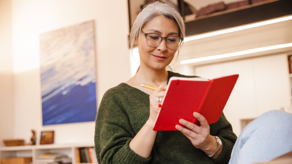 A woman wearing glasses is focused on writing in a red book, surrounded by a cozy, well-lit environment.