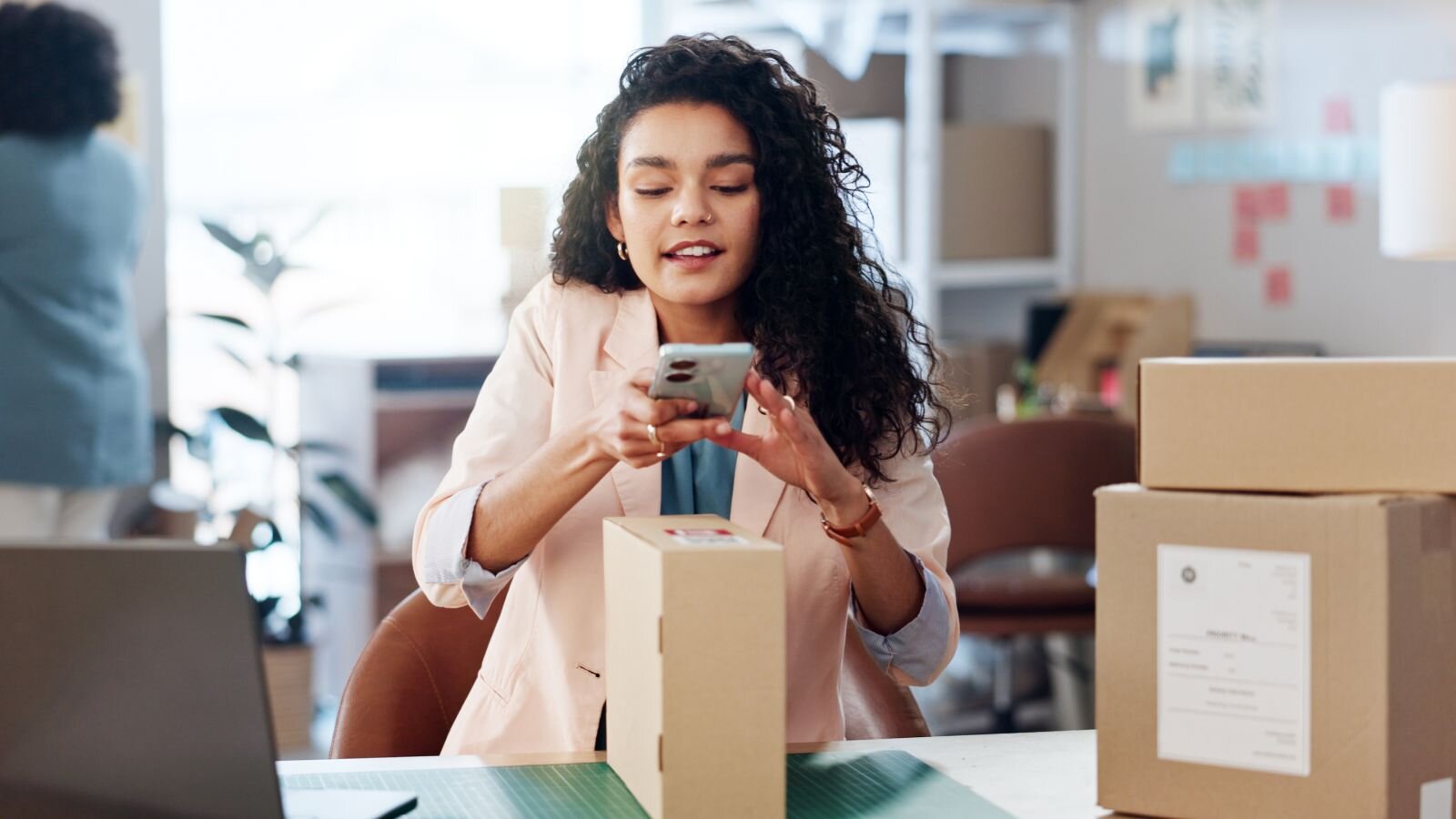 A woman in a light blazer uses her smartphone to scan a package in a modern office with cardboard boxes in the background.