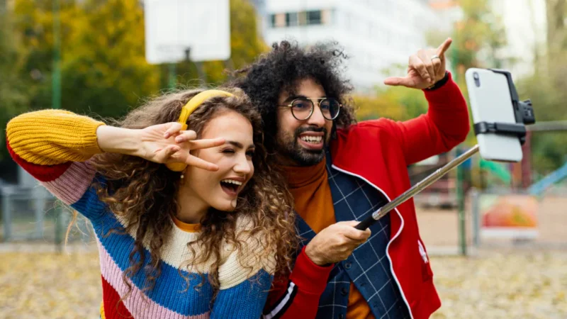 A joyful young couple in colorful sweaters takes a selfie with a phone on a selfie stick, both smiling widely.