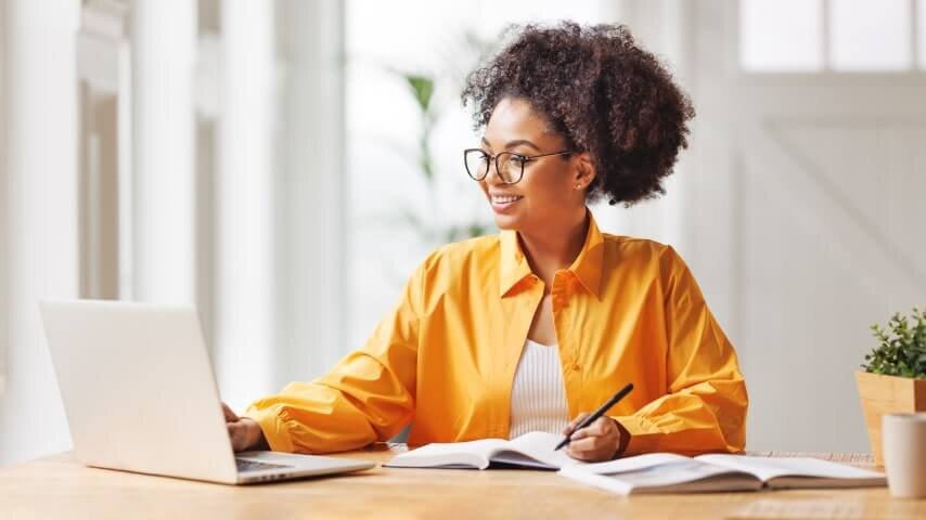 A smiling woman with glasses working on her laptop and taking notes.
