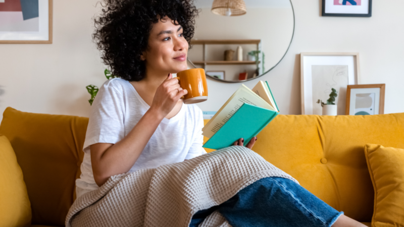 A woman with a blanket sits on a couch reading a book.