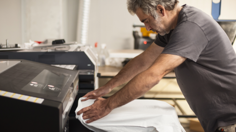 A man is working on an industrial printer, preparing an organic t-shirt for printing.