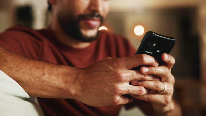 A man sitting on a couch is looking at his smartphone with a focused expression, reading about how to get more Etsy reviews.
