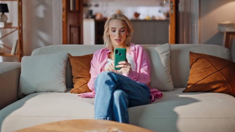 A woman in a pink shirt relaxes on a beige sofa, looking at her smartphone. With brown pillows on the sofa and a blurred kitchen background.