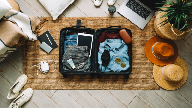 A travel bag full of clothing surrounded by different travel accessories (shoes, hats, laptop, charger).