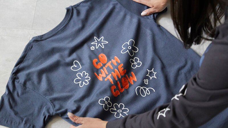 A person is putting a dark blue “Go with the glow” t-shirt on a surface.
