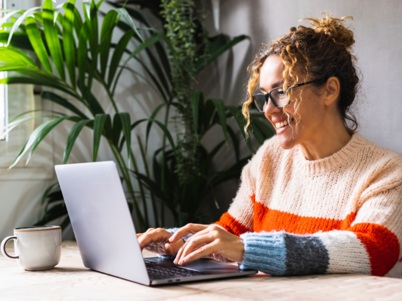 A woman in a colourful sweater smiles while using a laptop to create sticker designs.