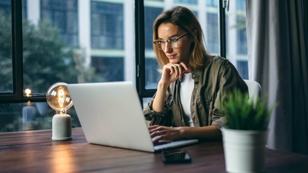 A woman wearing a dark green shirt, looking at a laptop screen and thinking.