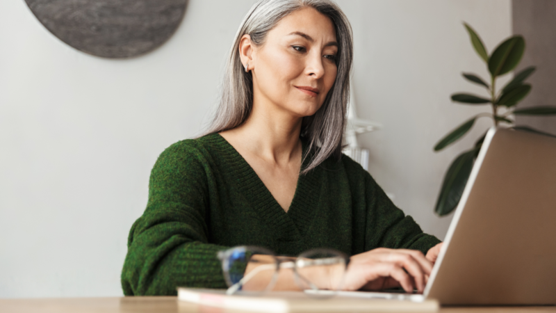 A woman with gray hair is sitting at a desk, focused on a laptop. She is wearing a green sweater, with glasses resting on the table.