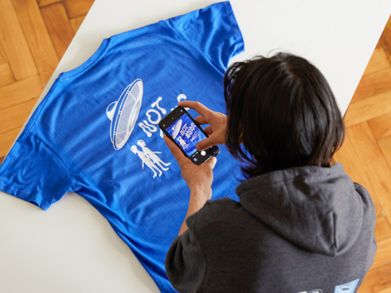 A man in a hoodie photographs a blue t-shirt with a UFO and aliens graphic, and the text "Not alone".