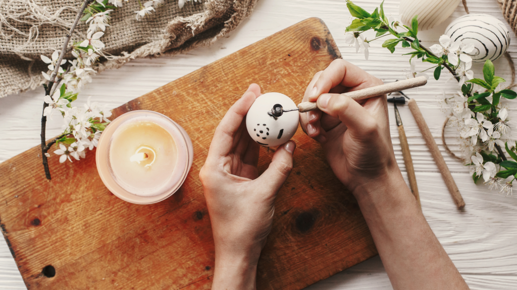 Hands delicately decorate an egg with a wax tool on a wooden board. A lit candle and cherry blossoms are nearby.