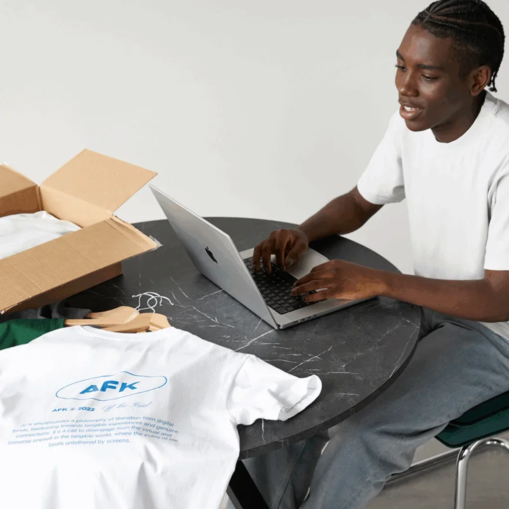 A man is sitting at a round table with a laptop and a white “AFK” t-shirt next to it.
