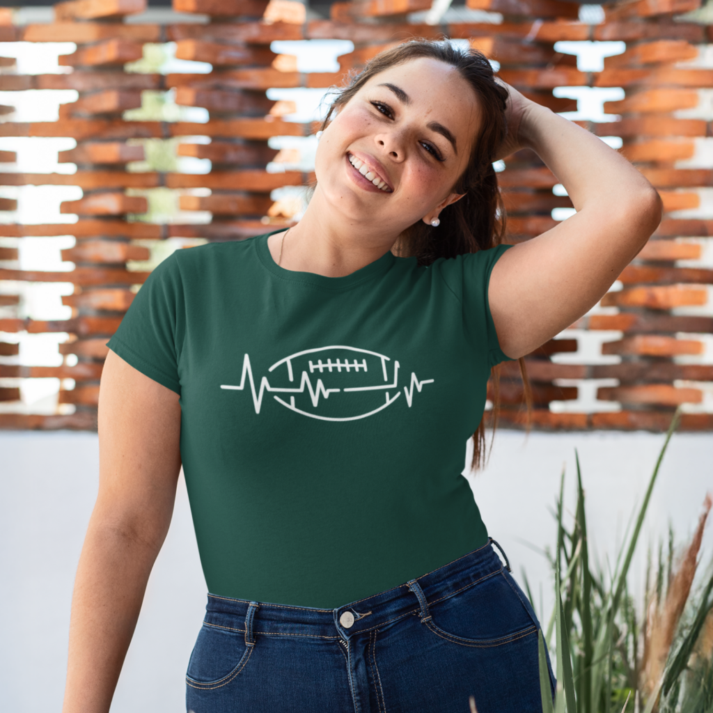 A smiling woman in a green shirt with a white heartbeat and football design, standing outdoors with a brick wall background.