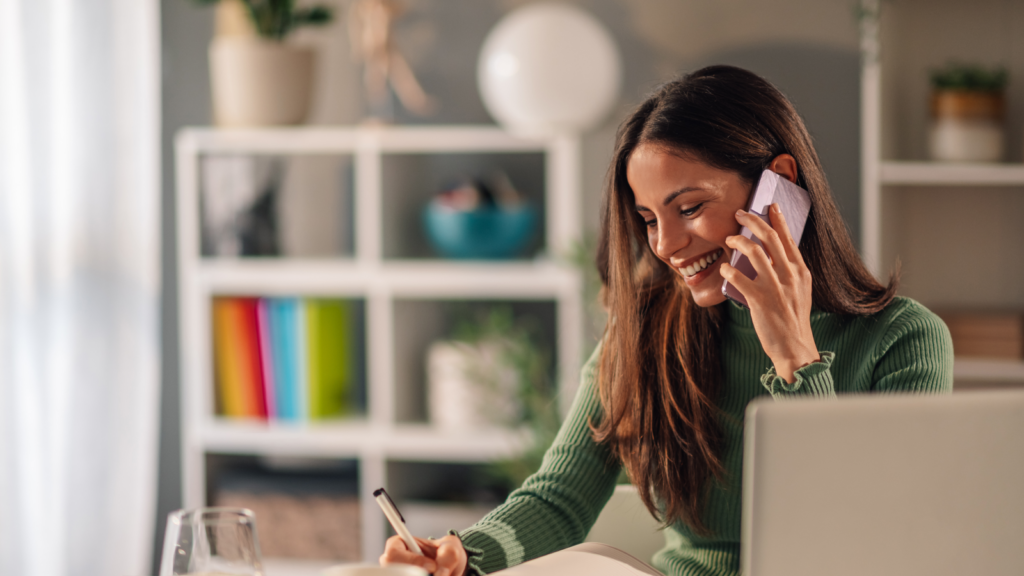 A smiling woman talking on a mobile phone and writing something down in a notepad.