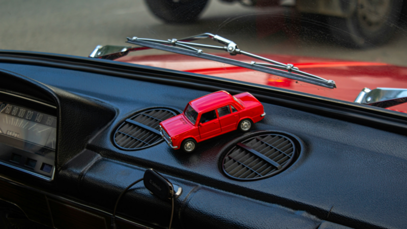 A red toy car is placed on a car dashboard, showcasing its vibrant color against the interior setting.