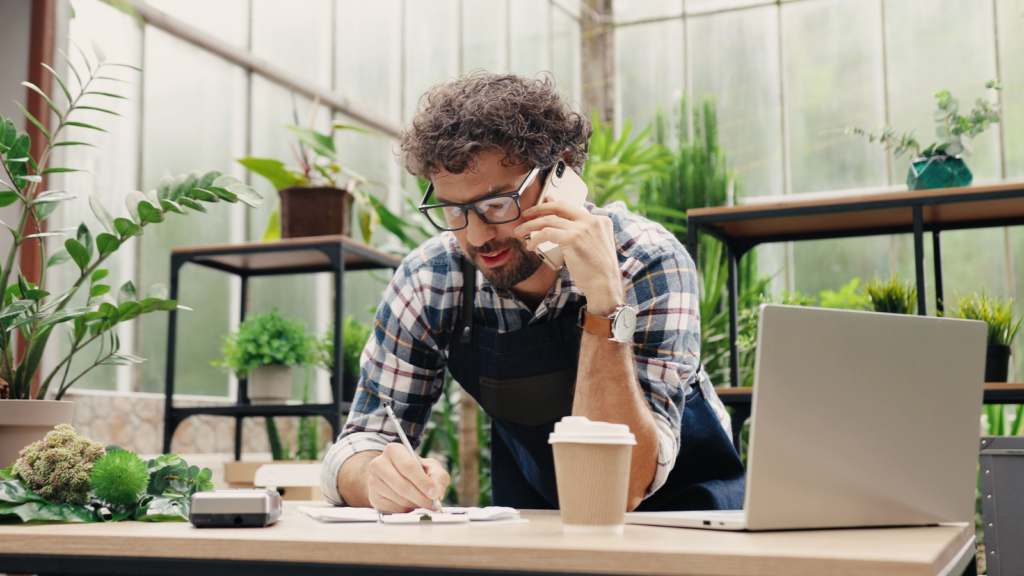 A man in an apron works on his laptop while speaking on the phone, balancing tasks in a busy environment.