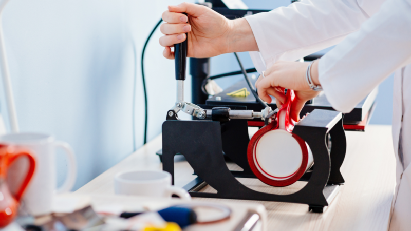 A person in a lab coat using a heat press machine to apply a design onto a mug.