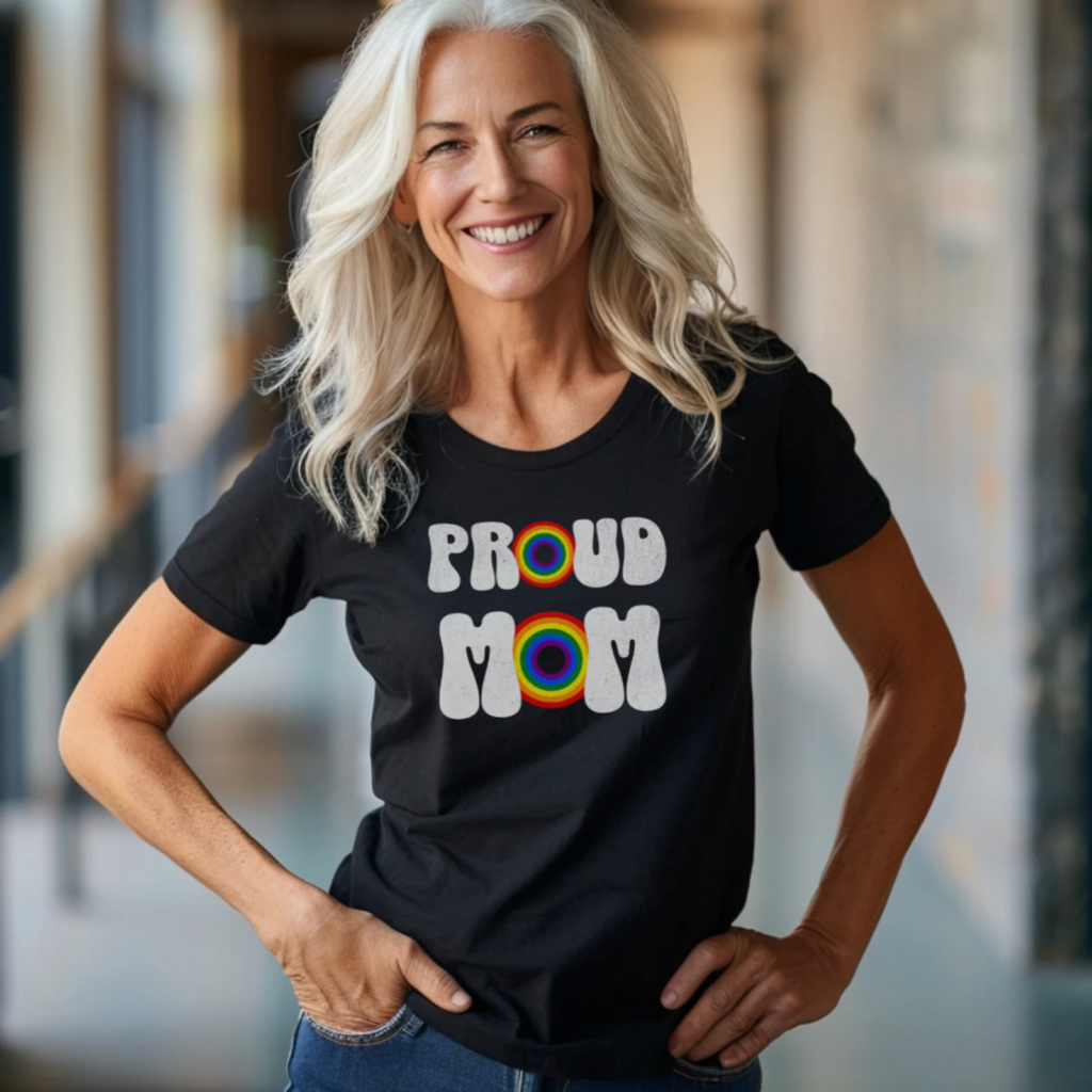Smiling woman with long silver hair wearing a "Proud Mom" t-shirt featuring rainbow letters.