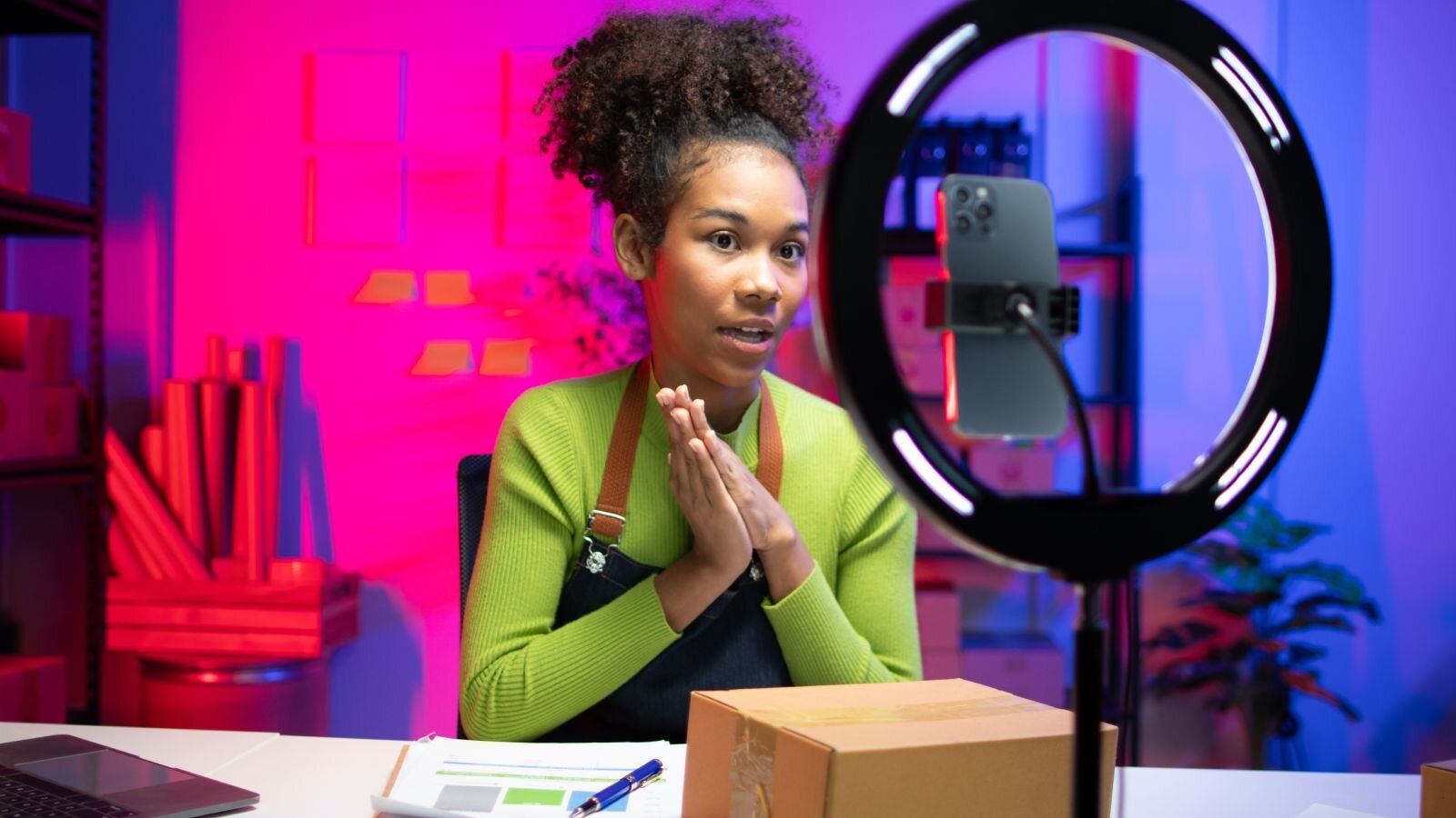 A woman wearing a green sweater and an apron, with curly hair, is sitting at a desk, recording herself using a smartphone.