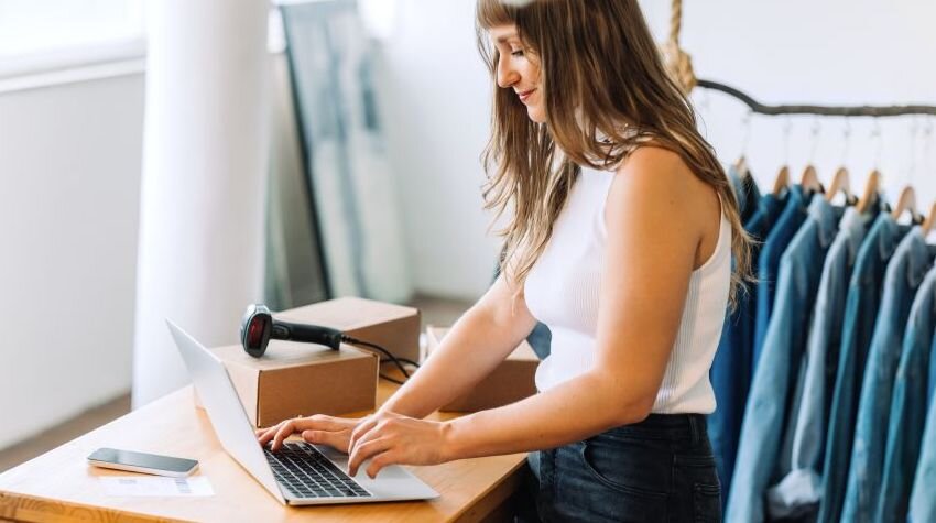 Woman working on her laptop, surrounded by clothing racks and dropshipping boxes.