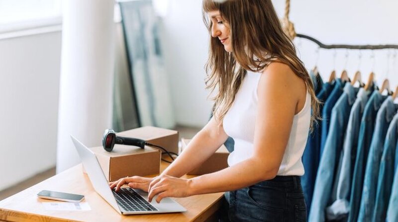 Woman working on her laptop, surrounded by clothing racks and dropshipping boxes.