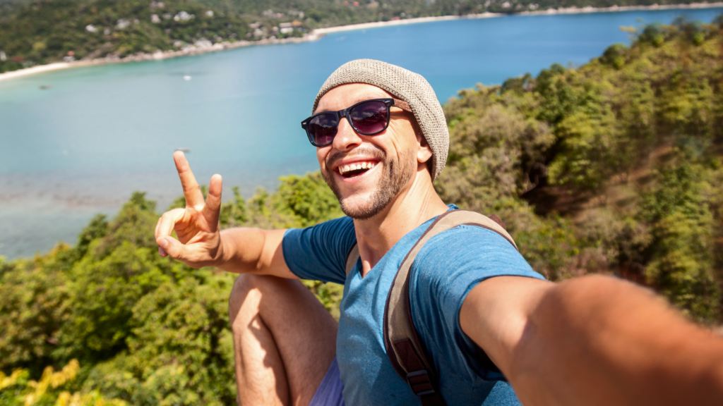 A man takes a joyful selfie overlooking a lush, green landscape and blue ocean. He wears sunglasses and a beanie.