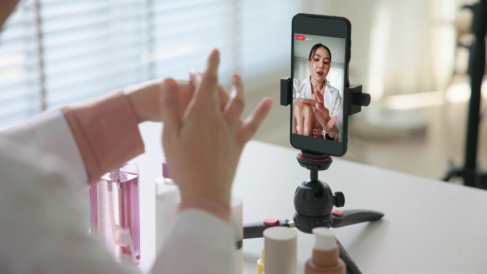 A woman in a lab coat demonstrates skincare on a live video stream, with various beauty products on a table.