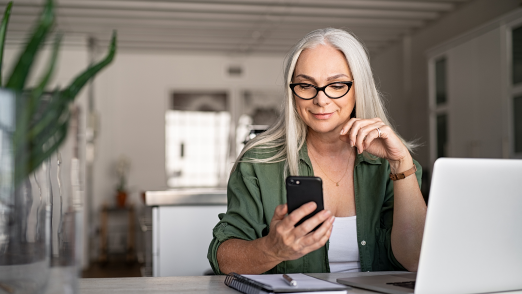 A smiling woman sitting behind a laptop and looking at a mobile phone screen, that she’s holding in her hand.