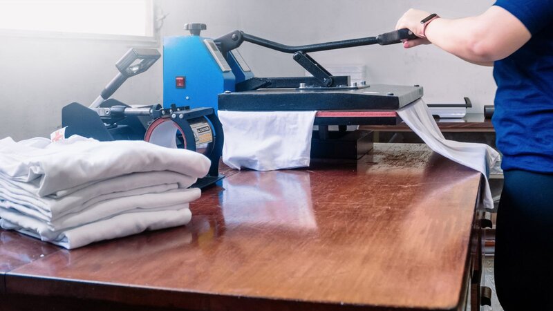 Close-up of a heat press machine printing a white t-shirt with folded shirts stacked nearby.
