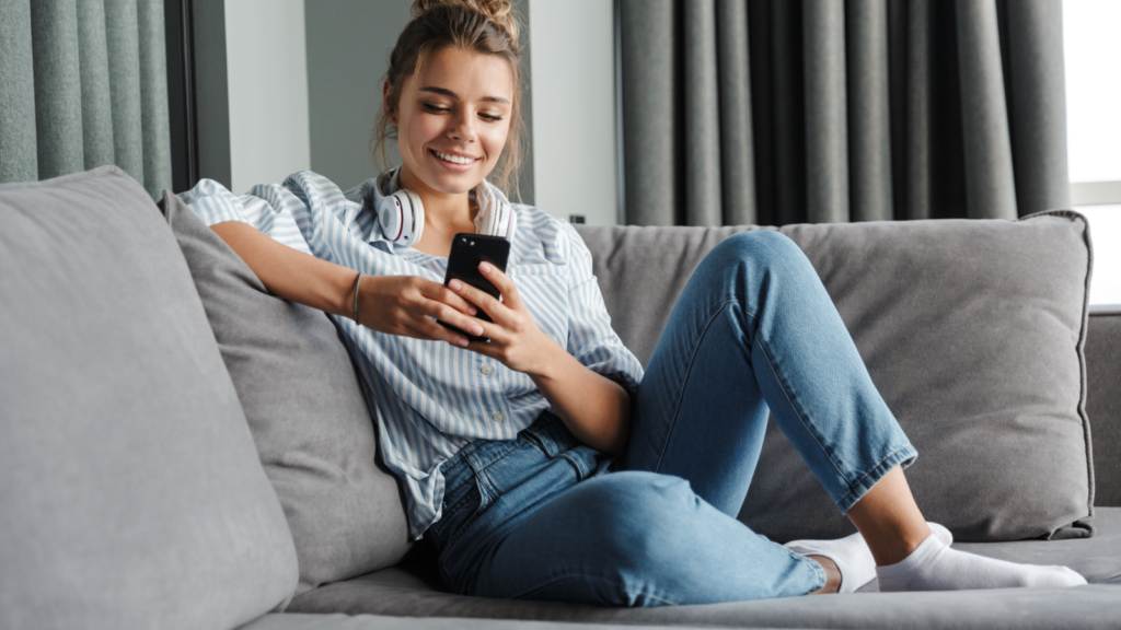 A smiling woman sitting on a couch and using a mobile phone.