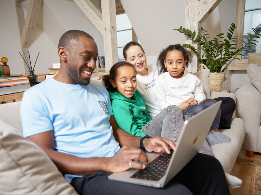 A family of four sitting on the couch and looking at something using a laptop.