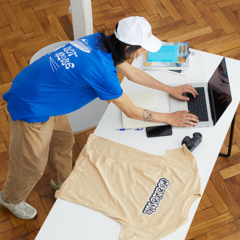Man in a blue shirt and white cap types on a laptop at a white table, surrounded by a notebook, phone, camera, and beige t-shirt.