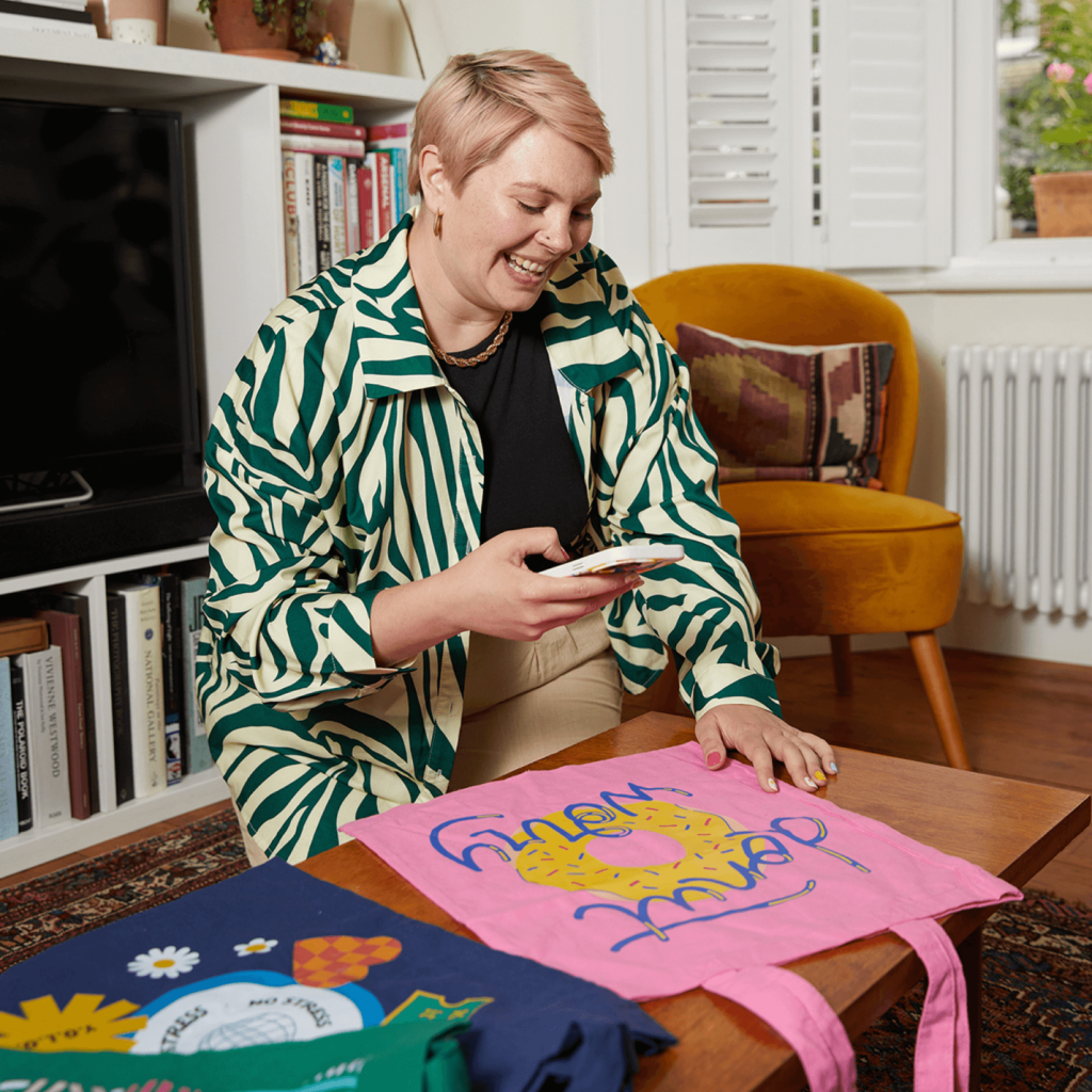 Smiling woman taking a photo of a custom tote bag, with a smartphone.