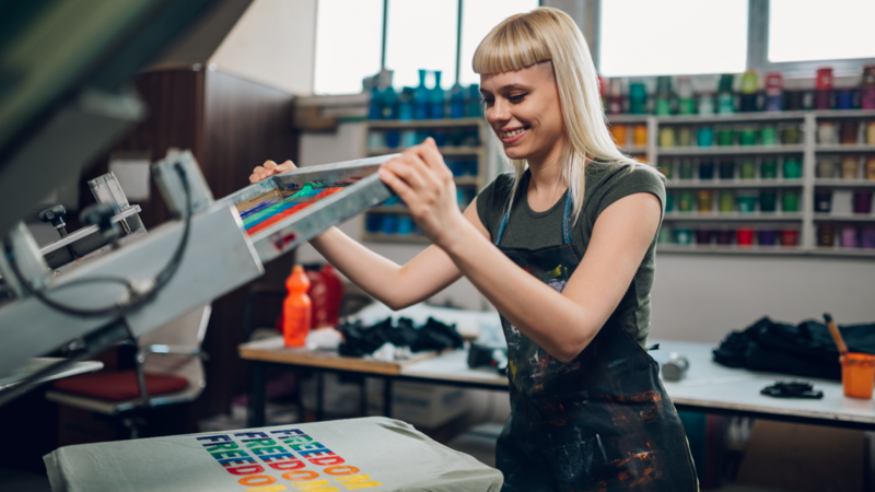 A smiling woman screen prints a colorful design on a T-shirt.