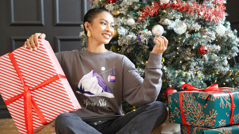 A woman in a gray sweater sits joyfully by a decorated Christmas tree holding a bauble. She is surrounded by red and green wrapped gifts.