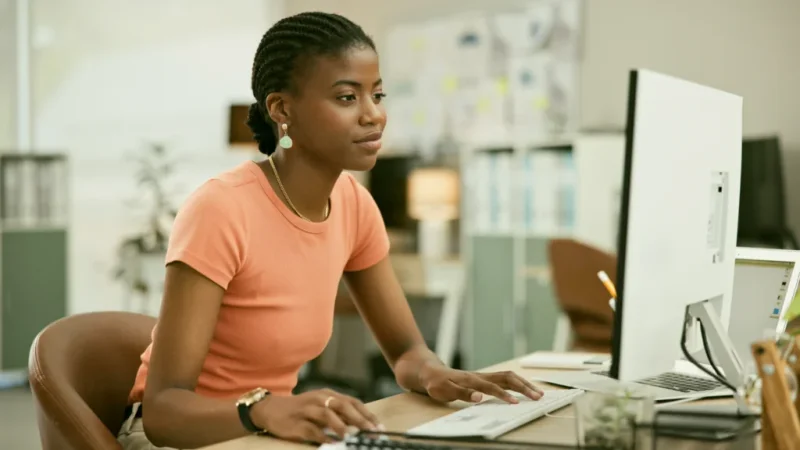 A woman in an orange shirt sits at a desk, focused on her work while using a computer.