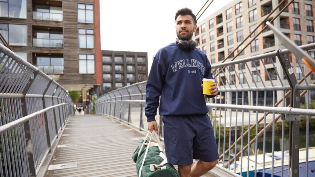 A man wearing custom sportswear, walking on a bridge while holding a bag and drinking coffee.