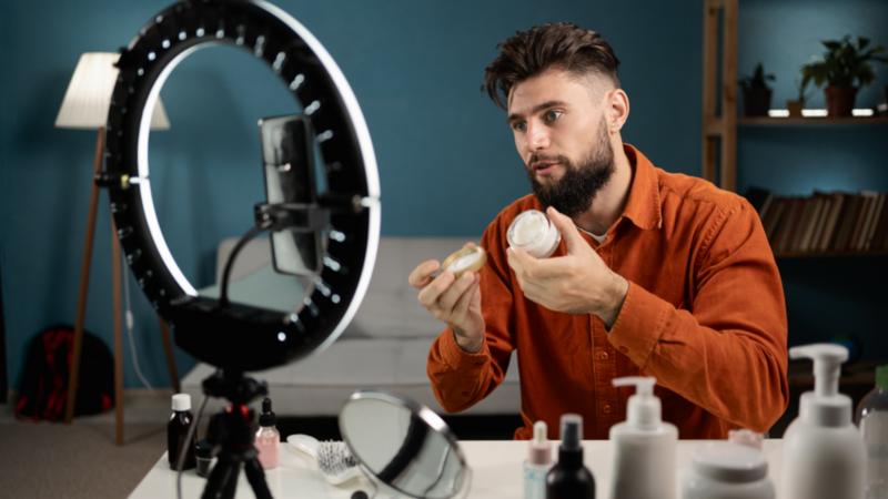 A man with a beard holds skincare products, demonstrating them in front of a smartphone and ring light.