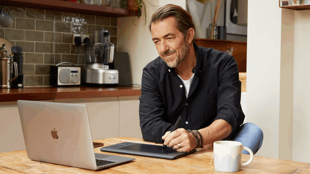 A man sitting in a kitchen and working using a laptop and a pad.