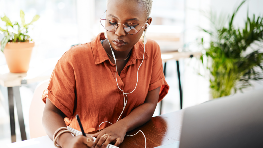 A woman in an orange shirt is engaged in writing, highlighting her concentration and creativity.