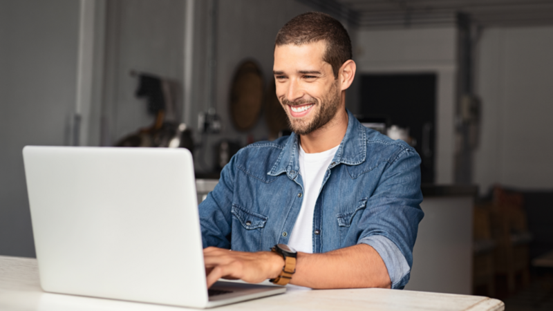 A smiling man is working on his laptop at an office desk, learning how to start a hoodie business.